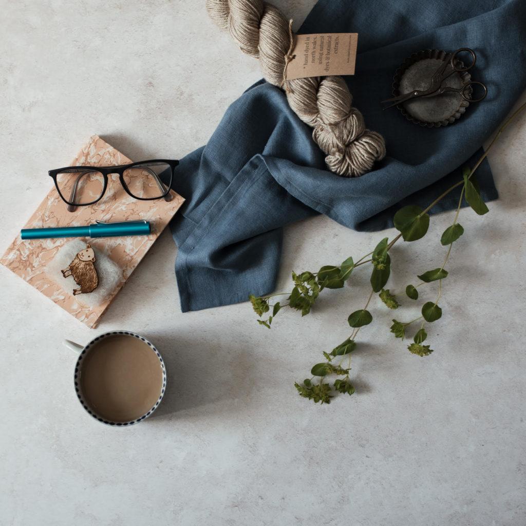Blue fabric with beige yarn, a cup of coffe scissors & spectacles with some greenery