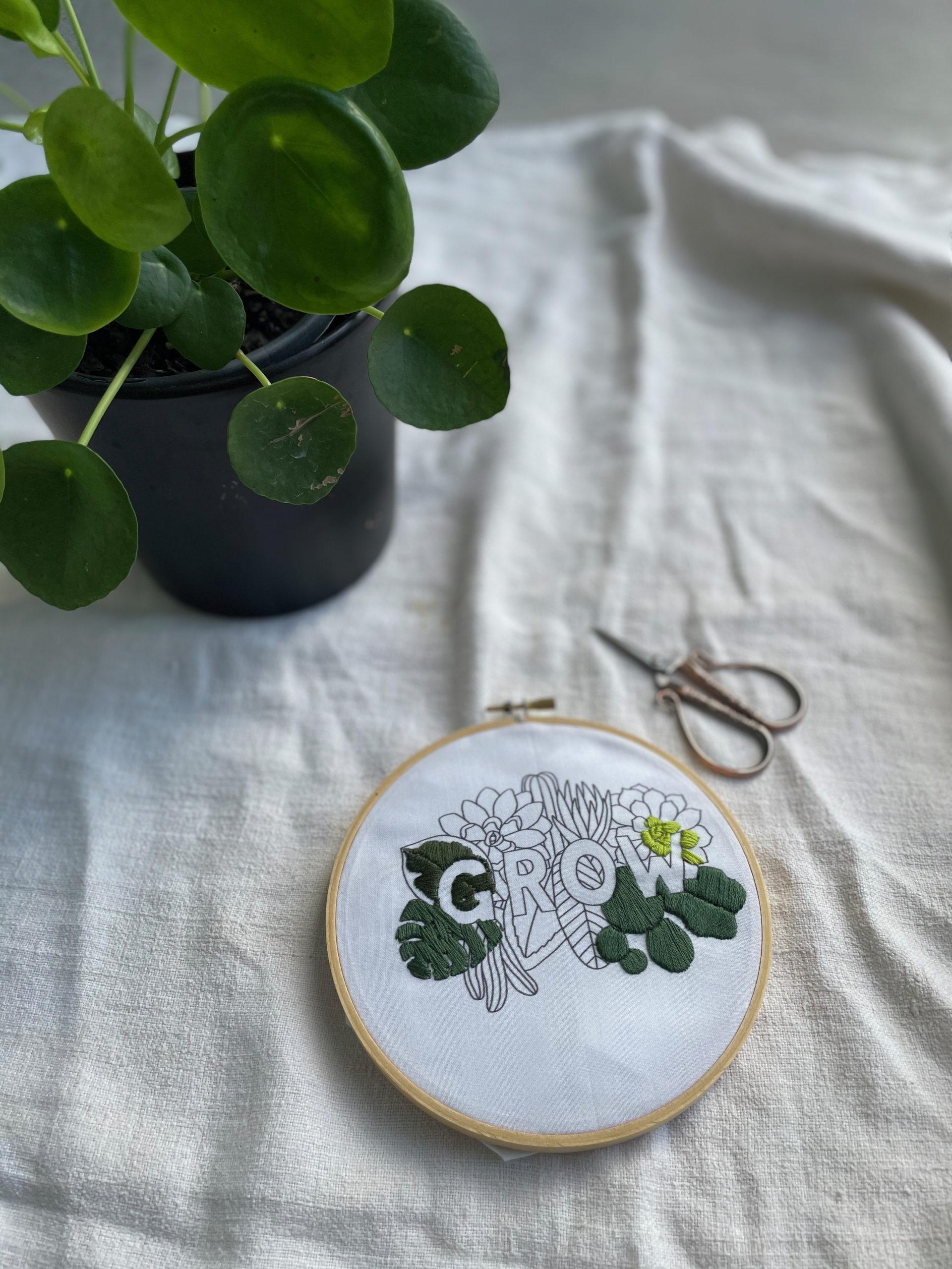 Embroidery with the word Grow, with green theads, in hoop. A plant and scissors are nearby