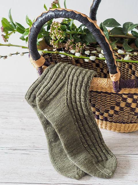 Curling Mist Socks by Curious Handmade made in a sage green yarn, resting on basket full of green tree cuttings