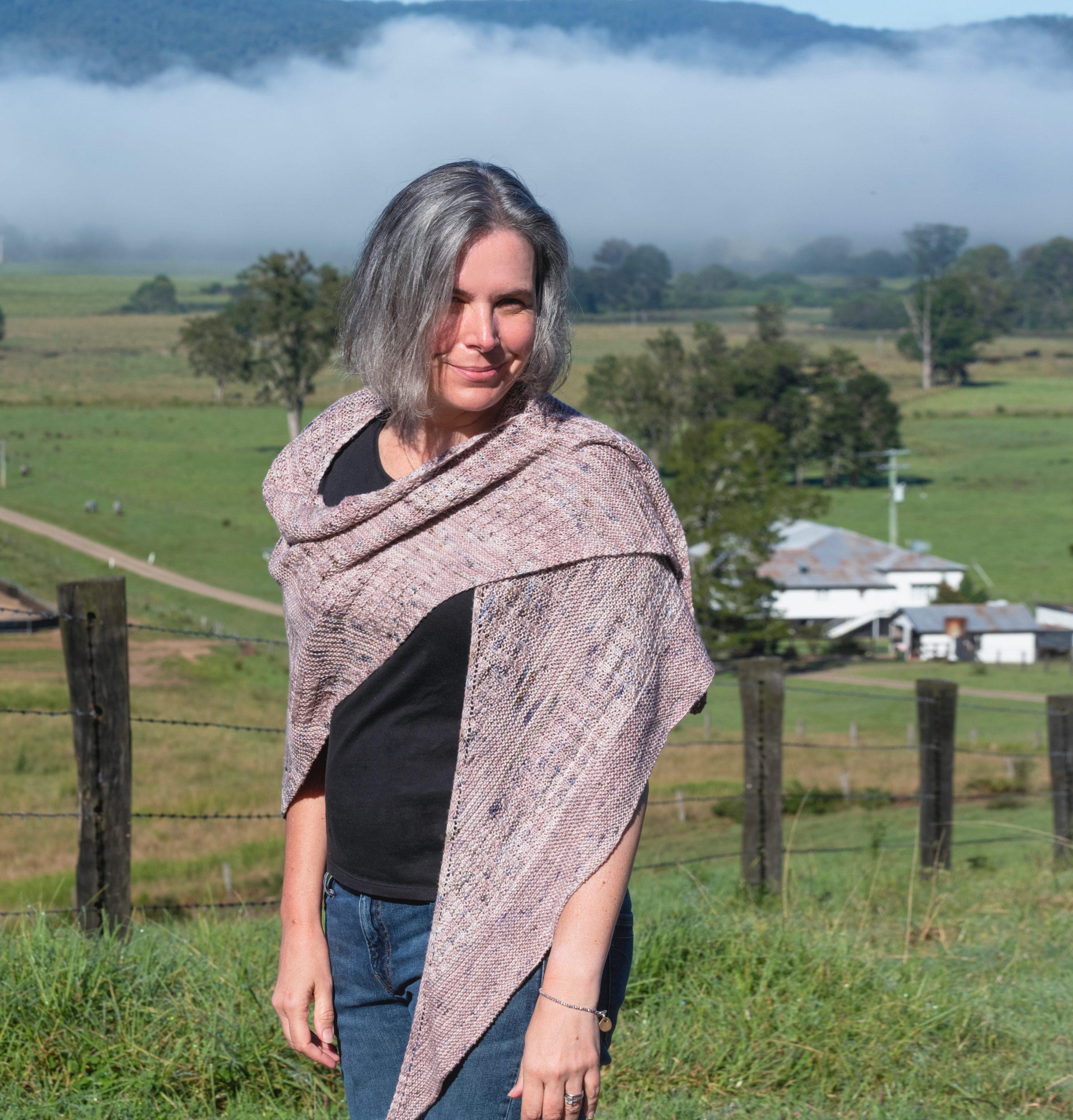 Helen Stewart, Curious Handmade wearing her own design, the Curling Mist shawl. There is green fields. and in the background, beyond the fields, a heavy curling mist rises into the hills