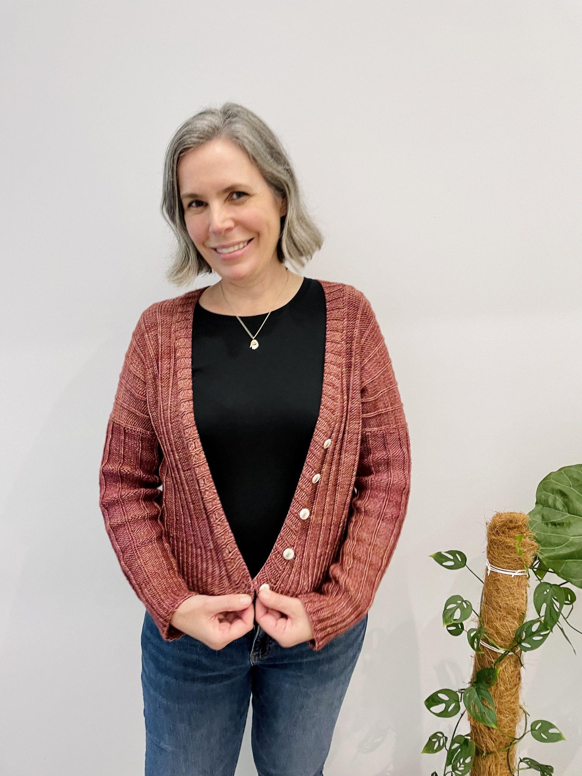 Helen Stewart, Curious Handmade, wearing a handknitted cardigan in a rose gold peach, design by Renee Callahan. There is a green climbing plant to Helen's right, and a white background