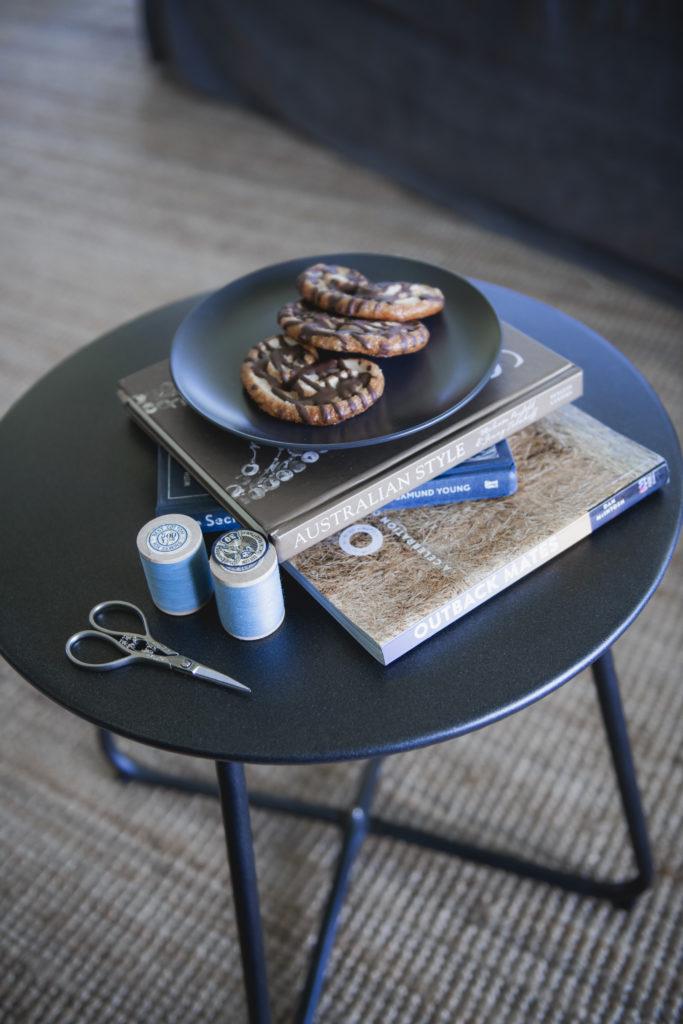 A plate of cookies on a black plate, sit on top of 3 books about Australia. Next to the books are two sewing reels with blue thread and a small scissors. All items are on a small round black table.