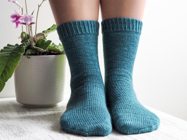 Feet wearing Helen Stewart's, Curious Handmade, design The Simply Curious Socks in a teal yarn, with a pink flower in a white flowerpot in the background