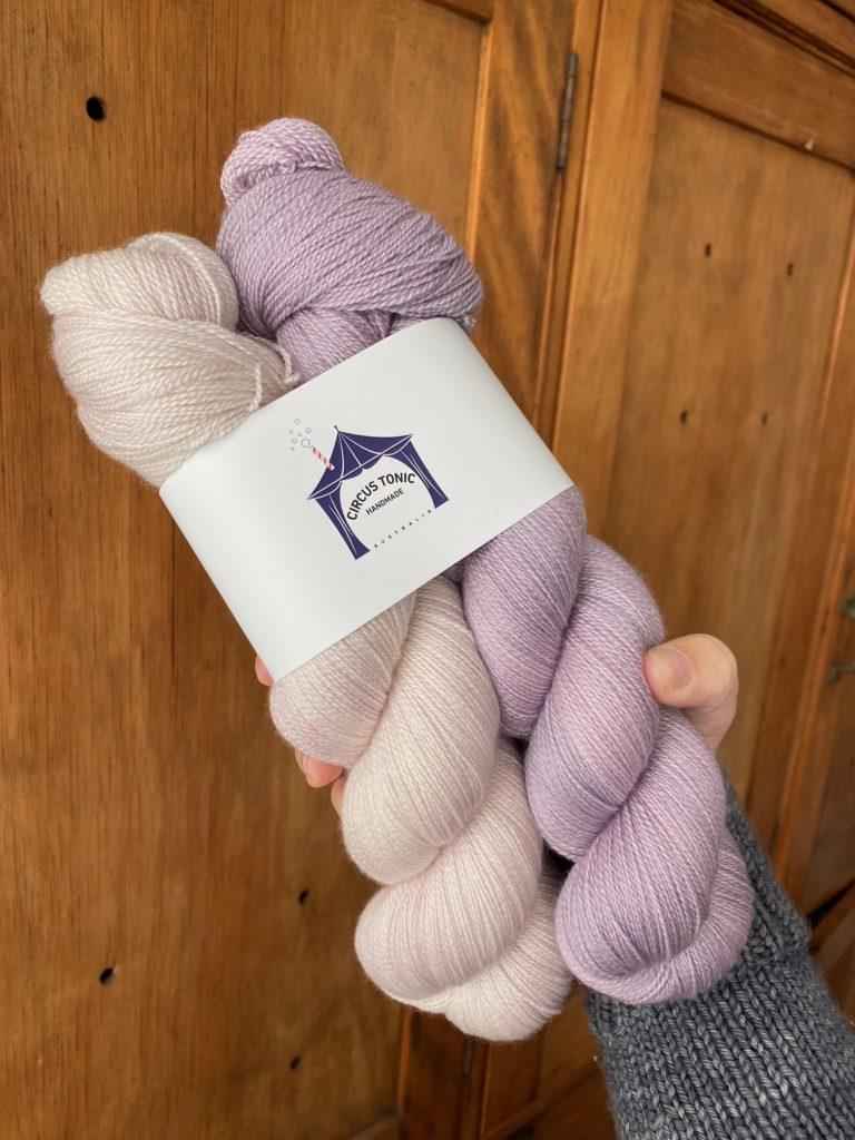 A hand holding a pale pink and pale purple skein of wool with the lable "Circus Tonic Handmade"