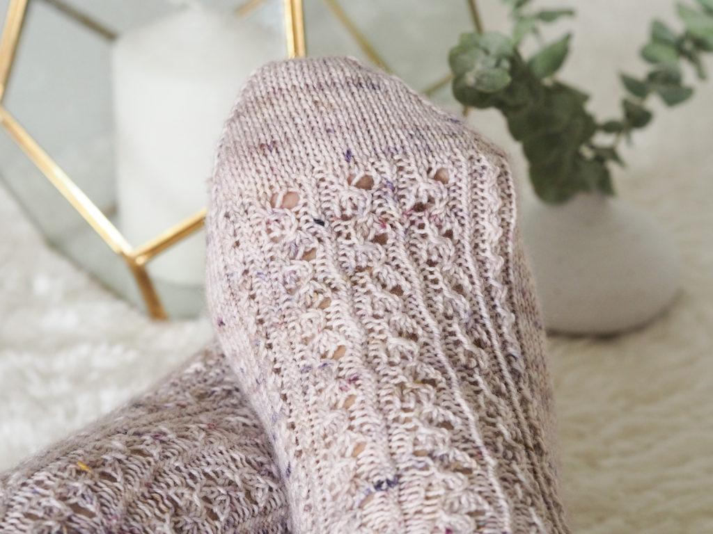 Feet wearing a pair of hand knit socks in a pale grey speckled yarn on a white table. In the background is a gold candle lantern and a little vase holding sprigs of eucalyptus