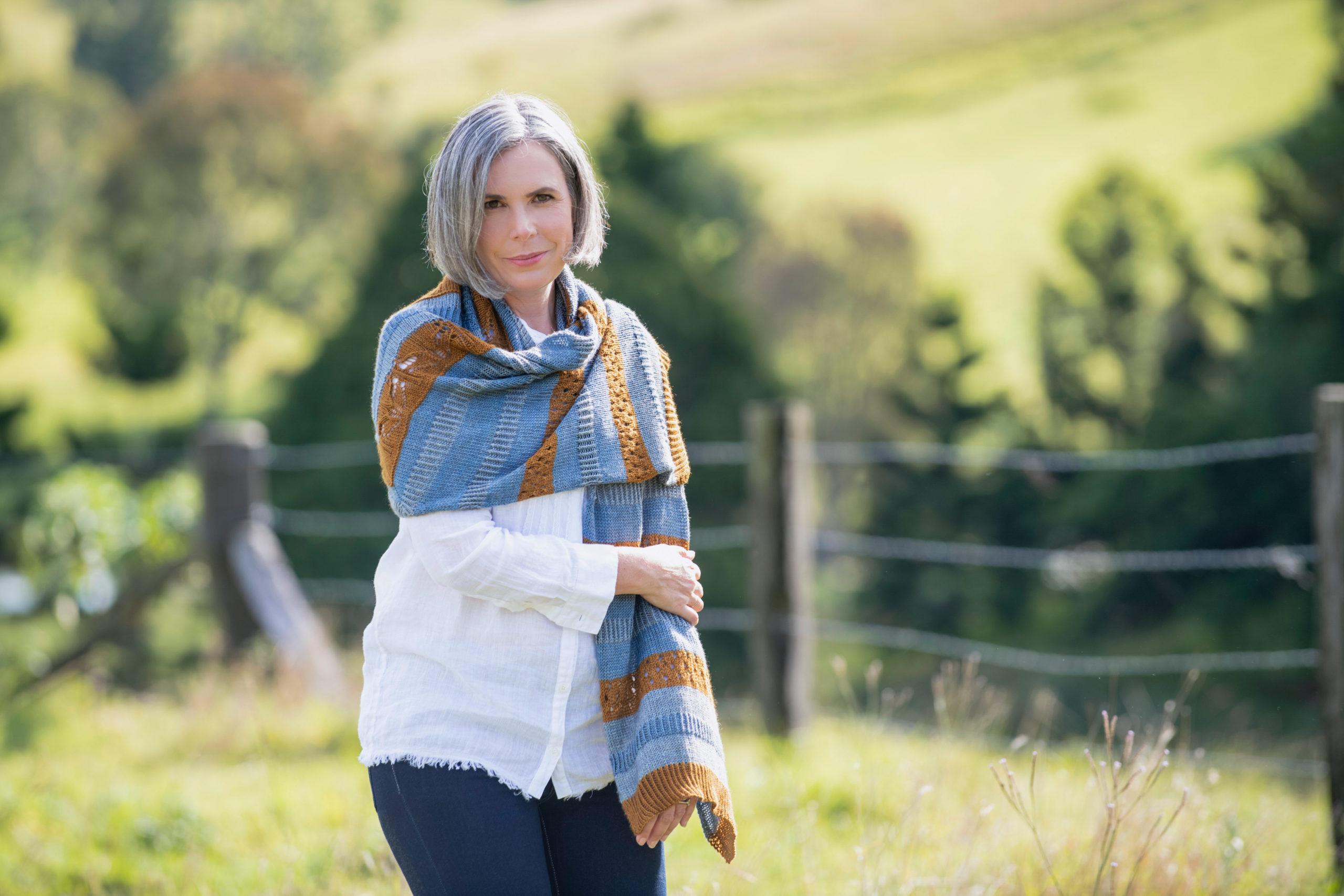 Helen stands in a green field, facing the camera wearing her own design The Wild Bees Wrap large rectangular hand knit wrap in blue, gold and grey yarns