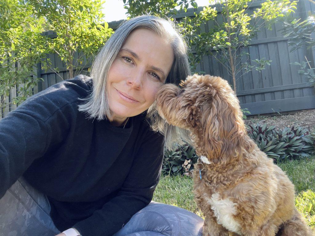 Helen Stewart, Curious Handmade in a garden with a fence behind her. Helen is crouching down and looking into the camera, and her golden dog Sindy is sniffing her face