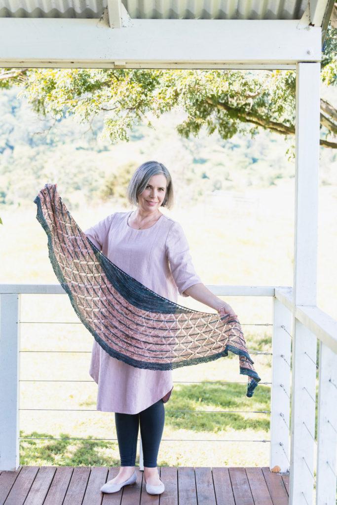 Helen Stewart, Curious Handmade, , wearing a light pink dress and leggings, stands of a white veranda. She's holding up a hand knit lace shawl, her own design The Veranda Shade Shawl in dark teal and pink speckled yarn