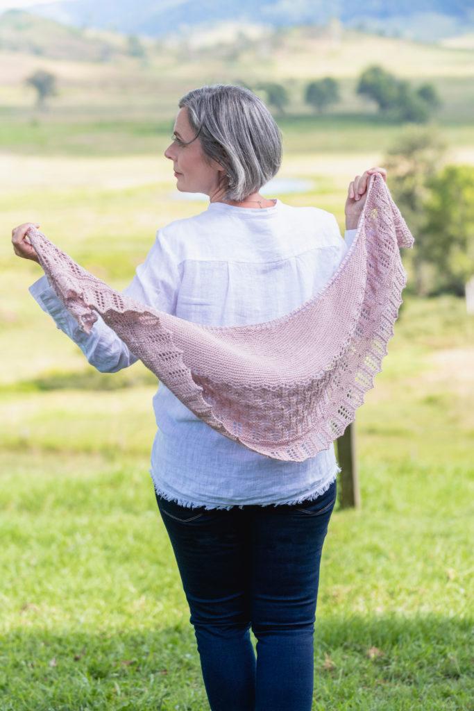 Helen walking in a field wearing a white linen top and dark blue jeans with her hand knitted pink shawl being pulled up to her shoulders. The shawl pattern is named the Blossom Time Shawl.