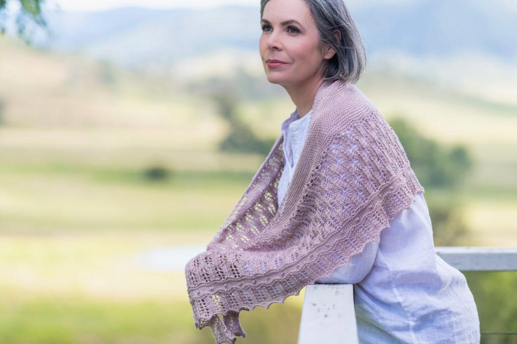 Helen, Curious Handmade, wearing a pale green top with a soft pink hand knit shawl around her shoulders, her own design the Blossom Time Shawl leans on a white fence.