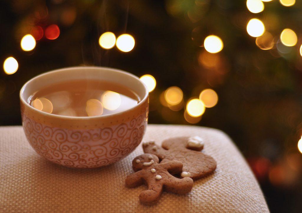 A cup of tea in a china cup with red swirls and a gold rim. ther are 2 ginger breadmen next to it, both sitting on a arm rest with twinkling christmas lights in the background.