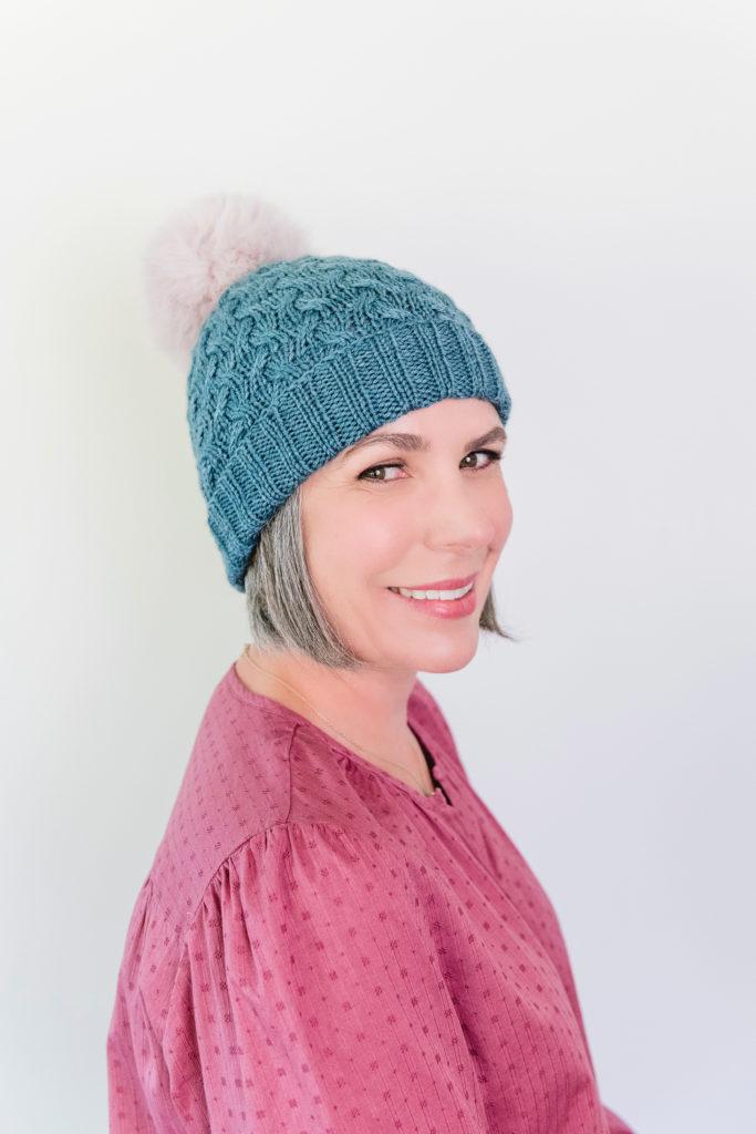 Helen wearing her newly knitted blue bobble hat with a white fluffy bobble on top. Helen is facing to the camera and smiling.