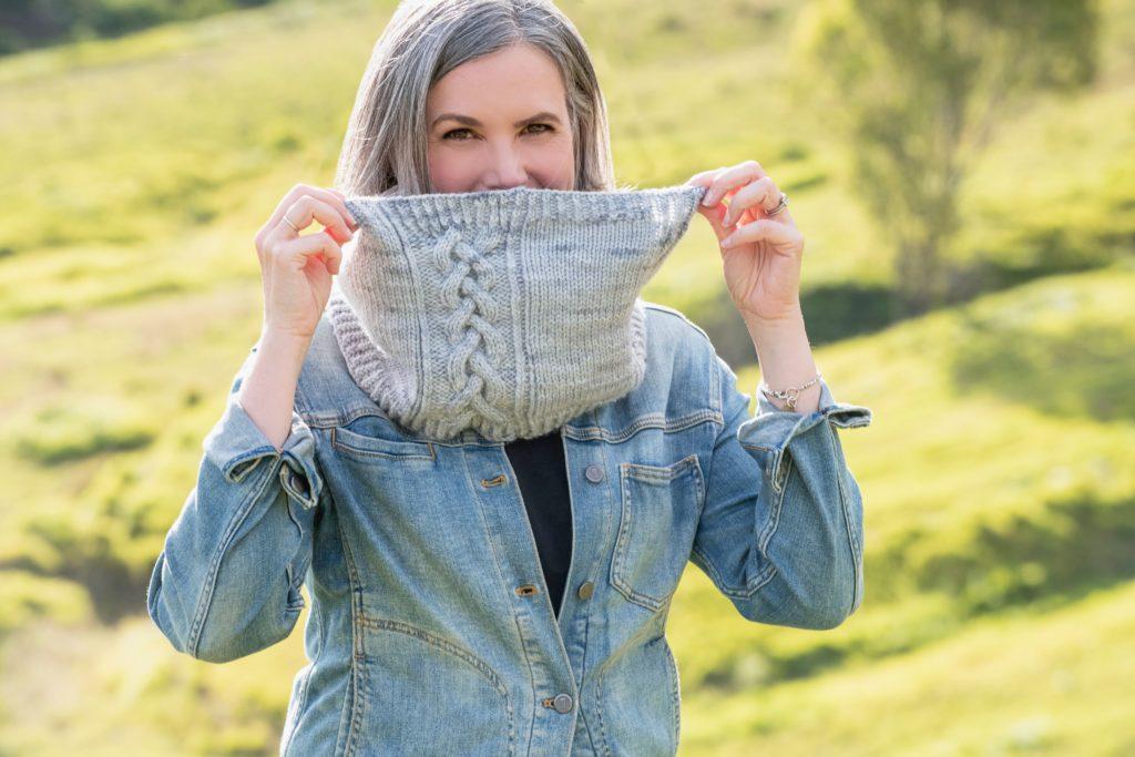 Helen from Curious Handmade, standing in the countryside wearing a light blue jean jacket, peeks over the top of a handknit cowl made of soft grey wool, featuring a braided cable design. Her own pattern called The Rose Cottage Cowl.