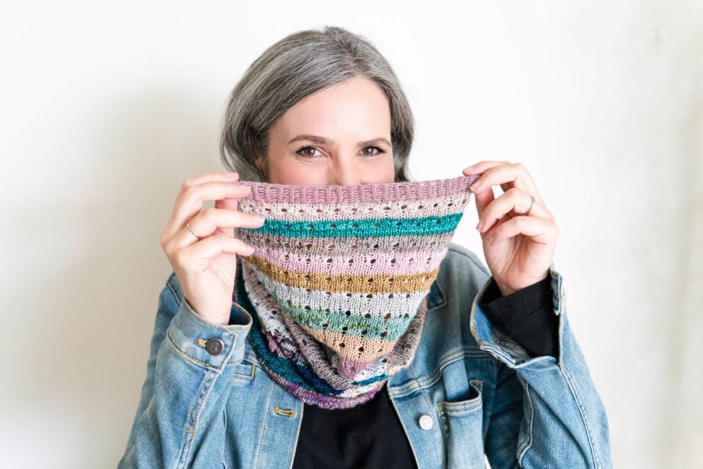 An image of Helen with her multi-coloured knitted cowl playfully covering half her face. Helen is smiling at the camera wearing a black top and light denim jacket.