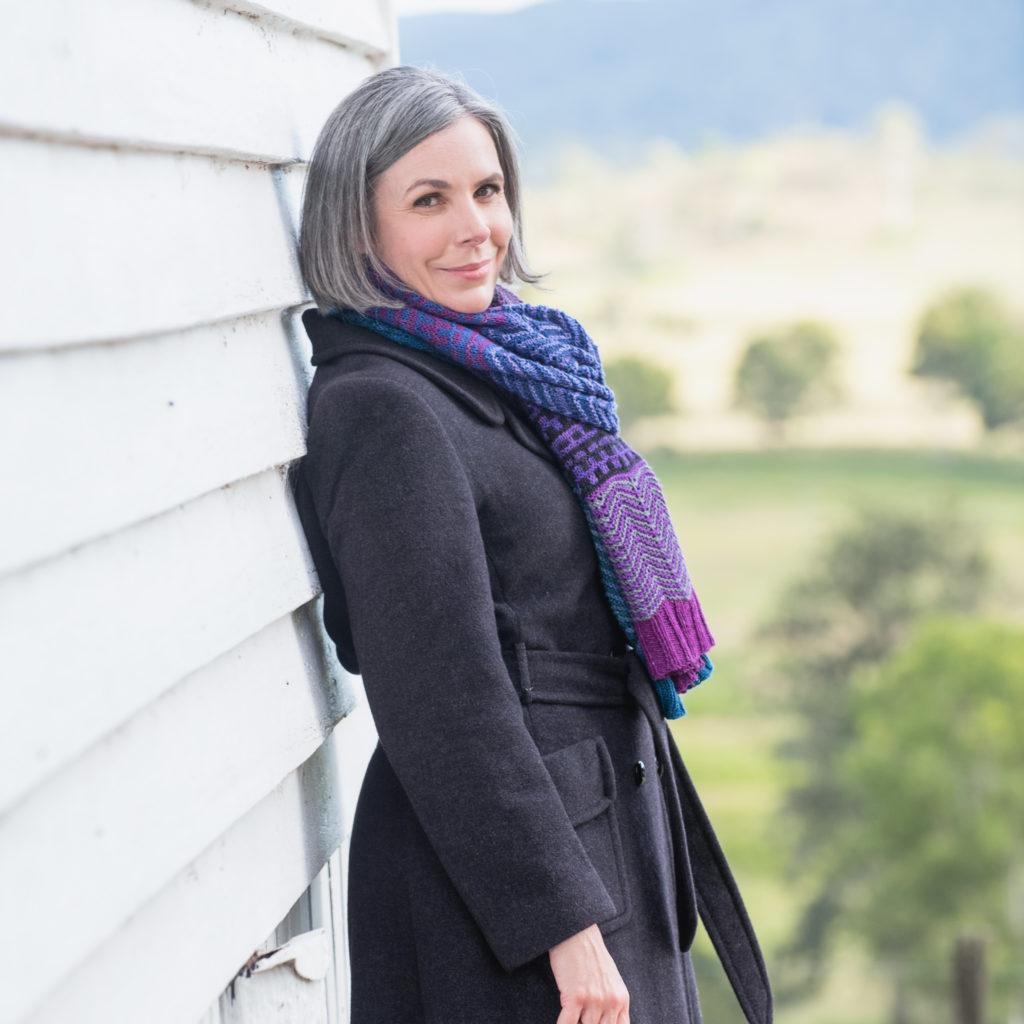 Helen wrapped in a warm wool jacket and a beautiful blue and purple hand knit scarf, leaning against a white slatted weatherboard house. Her own pattern called The Skyline Wrap.