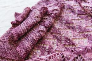 A close up shot of a handknit purple shawl, lying on a table. It has stripes of darker and lighter pink/purple yarn, with a zigzag lace pattern and tiny sparkling beads knit into the fabric.