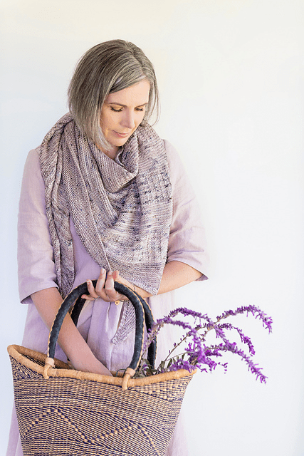 Helen Stewart wearing a handknit shawl featuring tiny cables, she's holding a straw basket with purple flowers