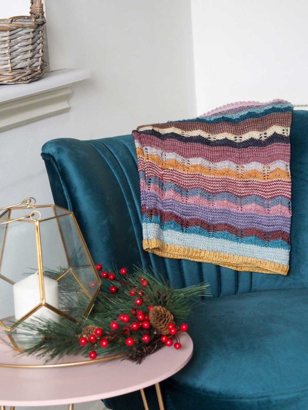 The Hearten Cowl is a multi-coloured striped extra long cowl on a blue velvet chair