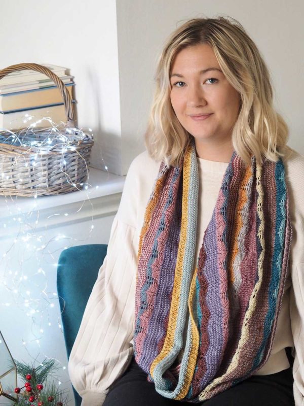 The Hearten Cowl is a multi-coloured striped extra long cowl laying on top of a finely knitted cream jumper