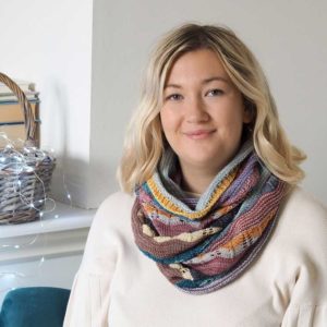 The Hearten Cowl is a multi-coloured striped extra long cowl wrapped around a womens neck and shoulders, on top of a finely knitted cream jumper