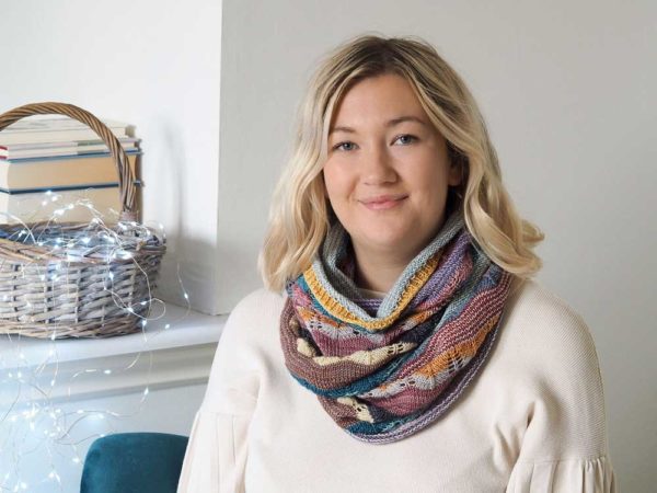 The Hearten Cowl is a multi-coloured striped extra long cowl wrapped around a womens neck and shoulders, on top of a finely knitted cream jumper