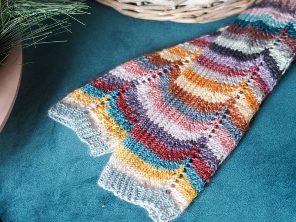 The Hearten Mitts are fingerless multi-coloured striped mitts next to a wicker basket