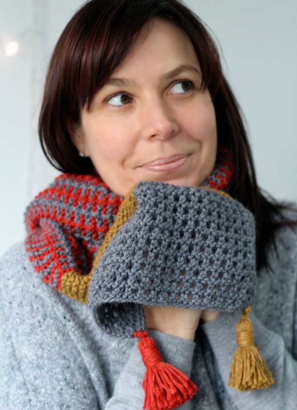 Helen wears a grey polo jumper with her Ice Skaing Scarf in red and grey checkered knit pattern, a think yellow stripe with a grey waffle-style knit at the end