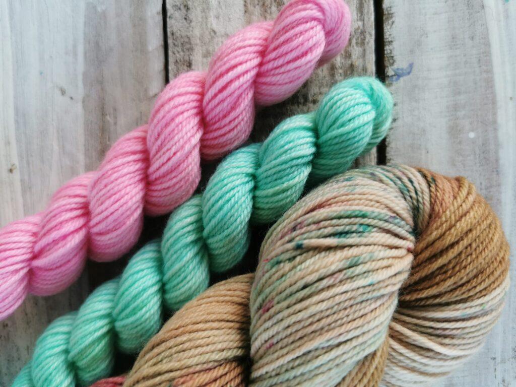 A pink mini skein of yarn, a teal mini skein of yarn and a speckled brown skein of yarn on a grey wood background