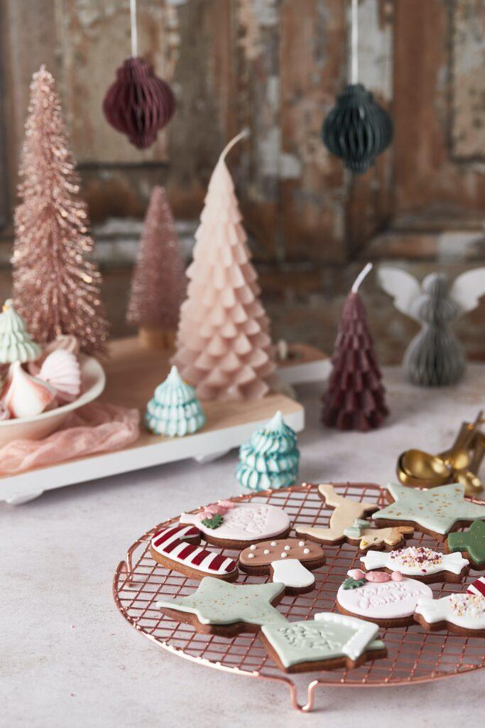 Miniature tinsel christmas trees and tree shaped candles in small colours sit behind a rack of decorated sugar cookies