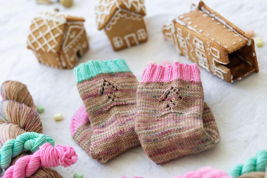 A pair of folded handknit socks with a gingerbread lace motif, sitting in between small gingerbread houses and skeins of yarn. 